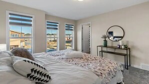 A bedroom with a bed, a bedside table with a mirror, and a view of buildings through large windows.