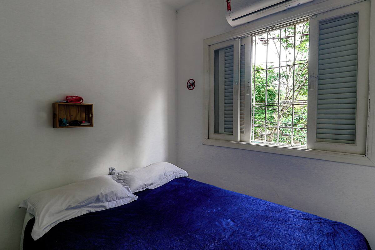Une chambre avec un lit simple, une fenêtre avec des volets, une petite étagère sur laquelle se trouve un objet rouge, et un panneau indiquant qu'il est interdit de fumer.