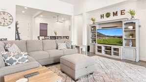 A modern living room with a sectional sofa, ottoman, and a flat-screen TV on a white entertainment unit. - Luxury Home in Litchfield Park w/Free Pool Heating (Litchfield Park)