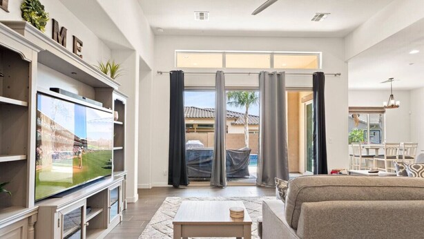 A spacious living room with a large flat-screen TV, a sofa, a coffee table, and a dining area visible through a glass door. - Luxury Home in Litchfield Park w/Free Pool Heating (Litchfield Park)