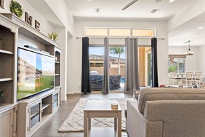 Ein großzügiges Wohnzimmer mit einem großen Flachbildfernseher, einer Couch, einem Couchtisch und einem Essbereich, der durch eine Glastür sichtbar ist.