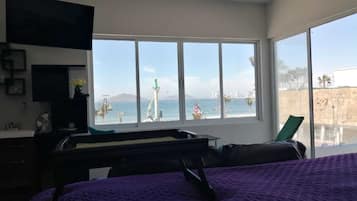 A bedroom with a large window, a bed, a desk, and a view of the sea and a lighthouse.