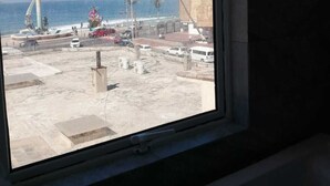Vista des d'una finestra sobre una platja, amb un far, palmeres i un paisatge urbà al fons.