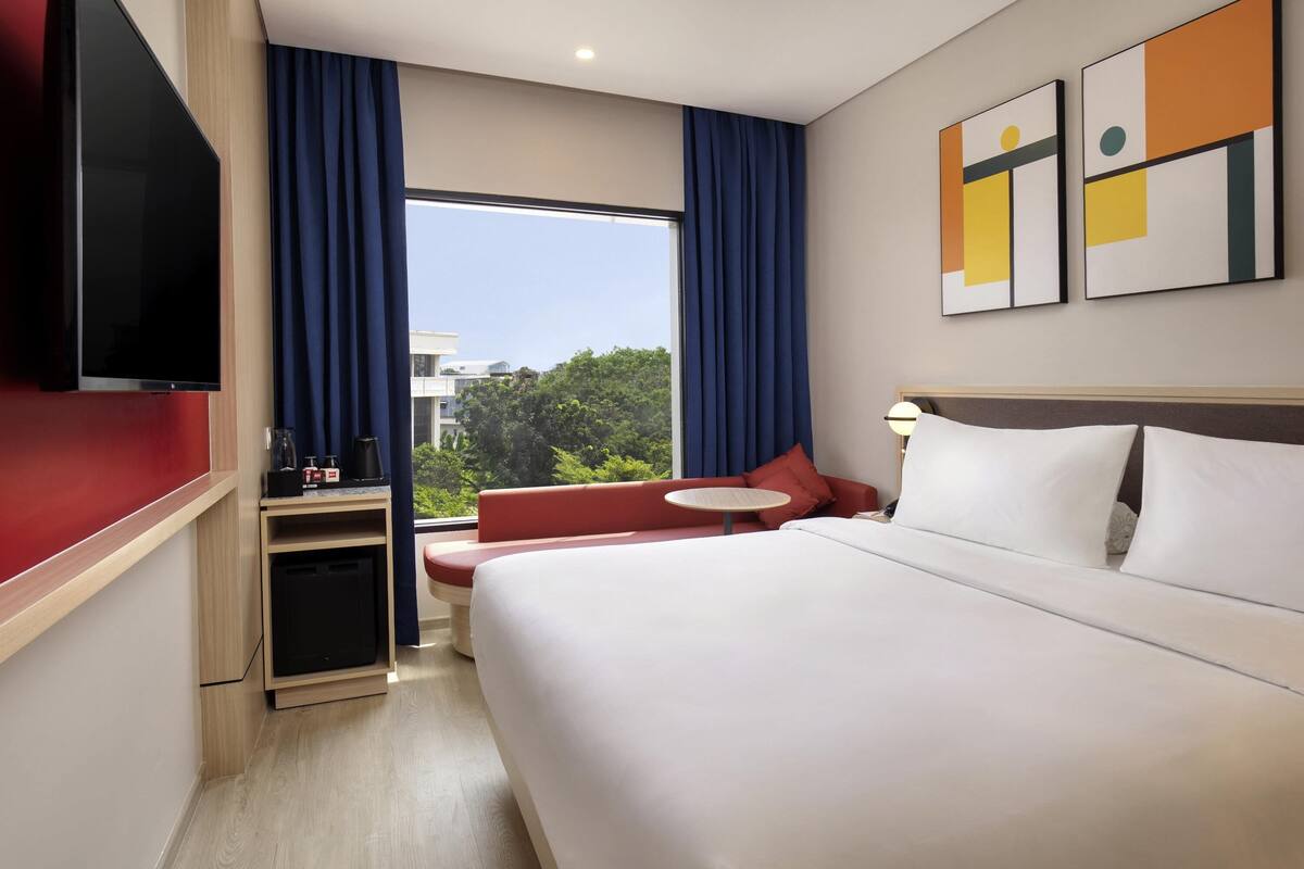 Quarto de hotel com cama, área de estar, televisão e janela com cortinas.