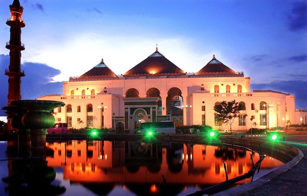 A large, illuminated building with three domed roofs and a central arched entrance, reflecting in a calm water body.