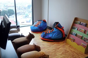 Zona de juegos infantil con vistas a la ciudad a través de un ventanal grande, un zapatero con zapatos de colores y una estantería con libros.