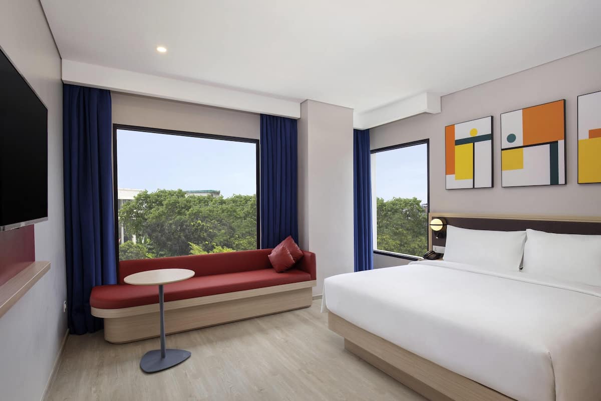 A hotel room with a large bed, a red sofa with a small table, and large windows with curtains.