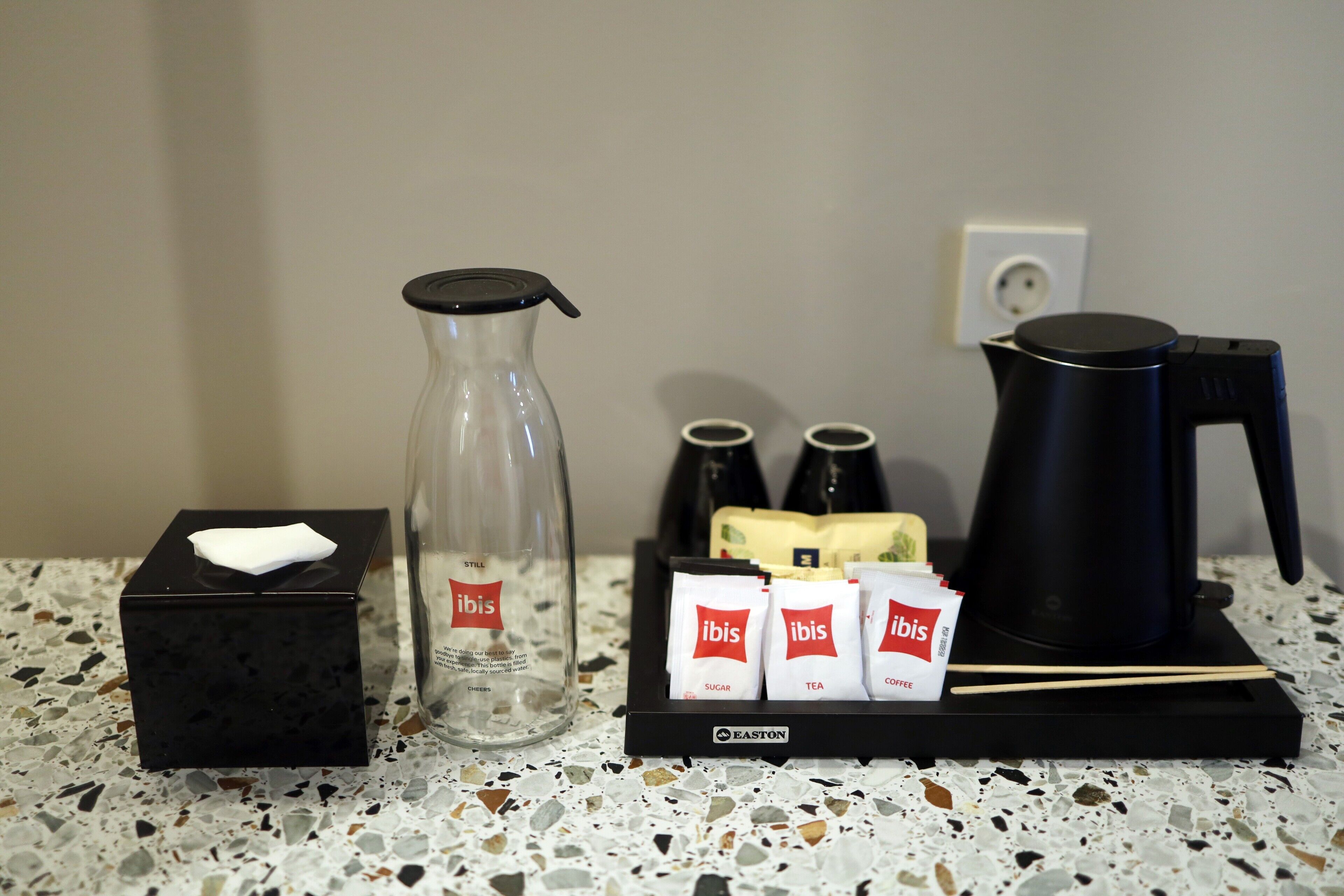 Un equipo de minibar con una caja de pañuelos, una botella de vidrio, una tetera eléctrica negra y tres sobres etiquetados 'ibis' para azúcar, té y café.