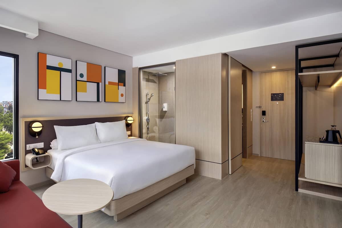 Quarto de hotel moderno com uma cama grande, mesa de cabeceira e uma pequena mesa redonda. Possui área de chuveiro com porta de vidro, armário e vista para o exterior.