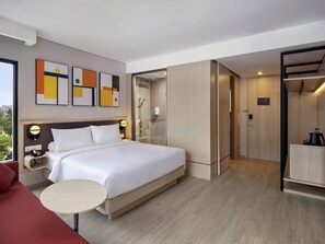 A modern hotel room with a large bed, bedside table, and a small round table. There is a shower area with a glass door, a wardrobe, and a view of the outside.