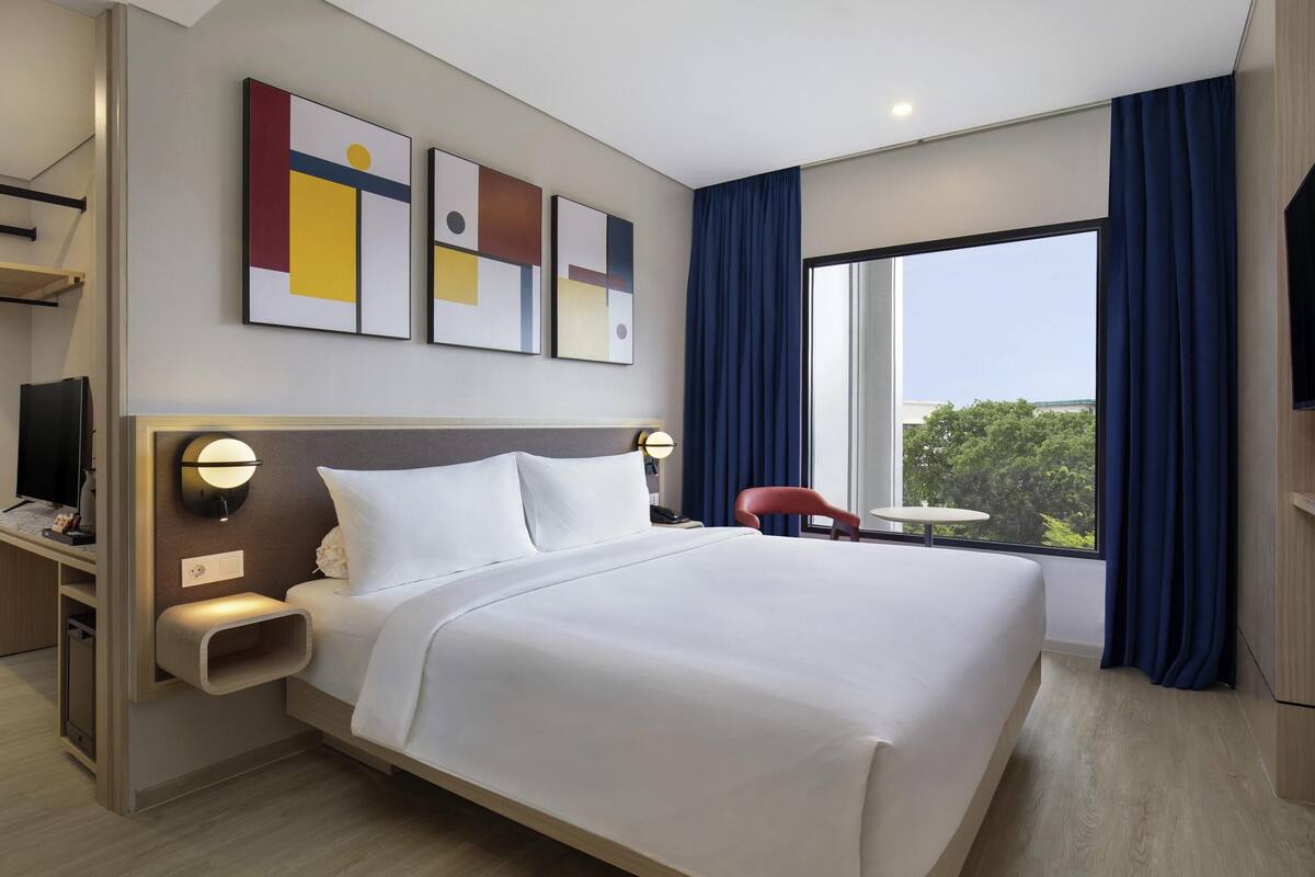 Quarto de hotel moderno com uma cama grande, uma mesa com televisão de tela plana, uma mesinha e vista para o verde lá fora.