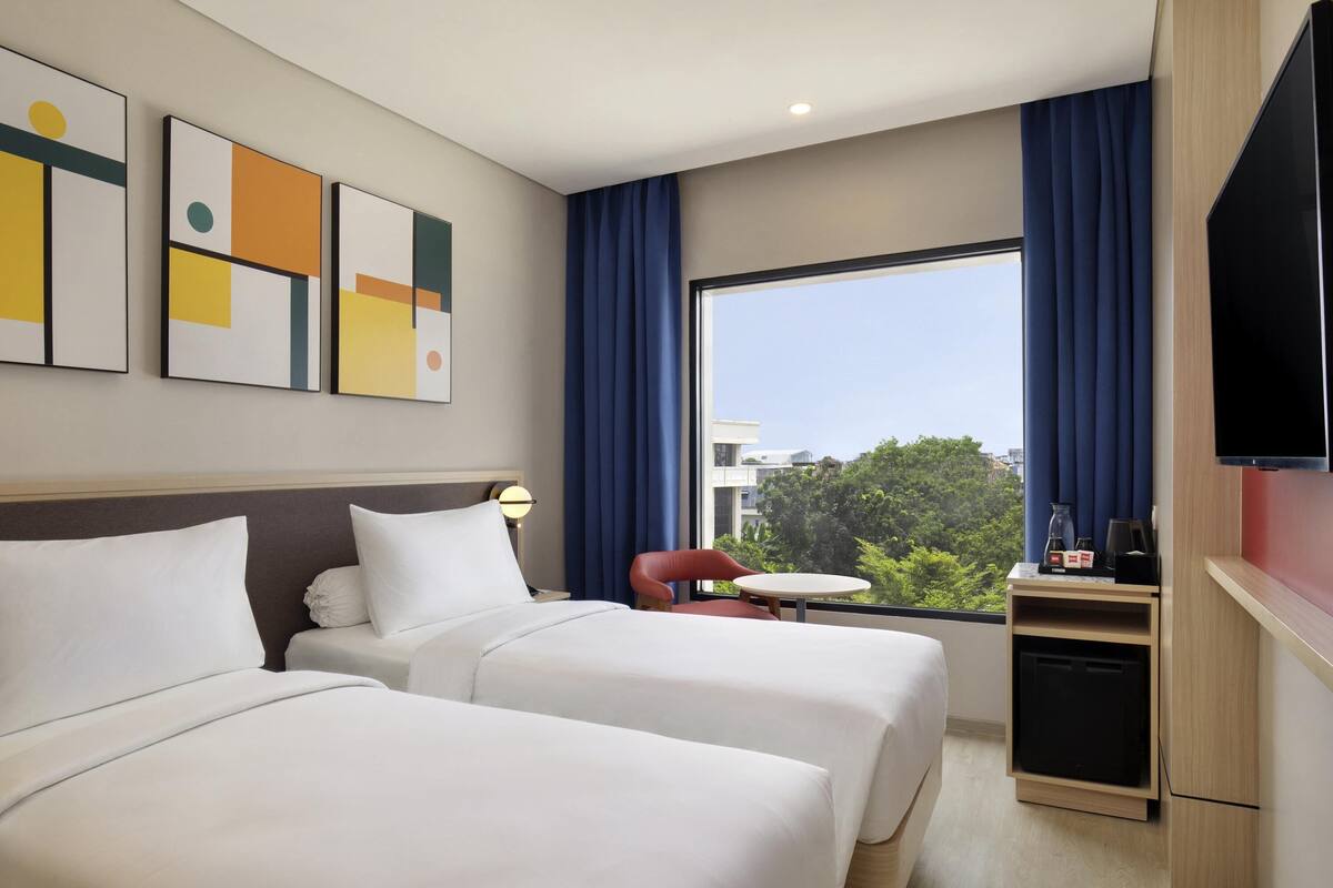 Quarto de hotel com duas camas, televisão, uma mesinha e janela com cortinas.