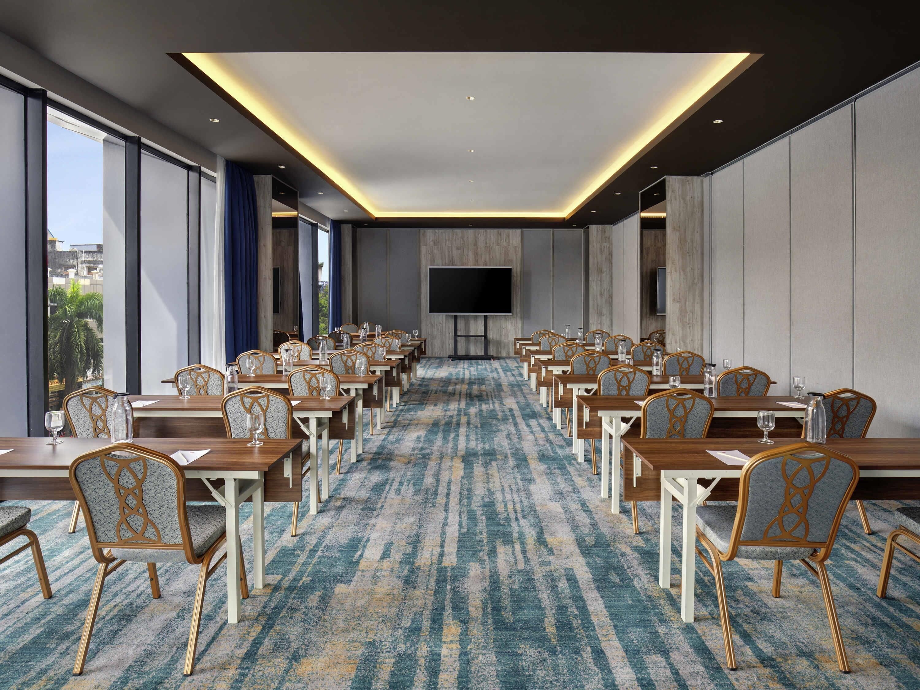 A conference room with rows of tables and chairs, a large flat-screen TV, and a patterned carpet.