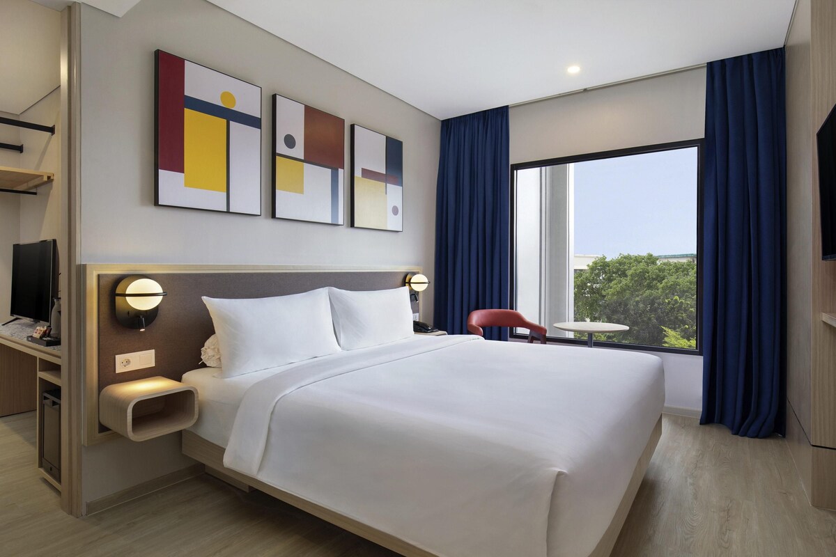 A modern hotel room with a large bed, a desk with a flat-screen TV, a small table, and a view of greenery outside.