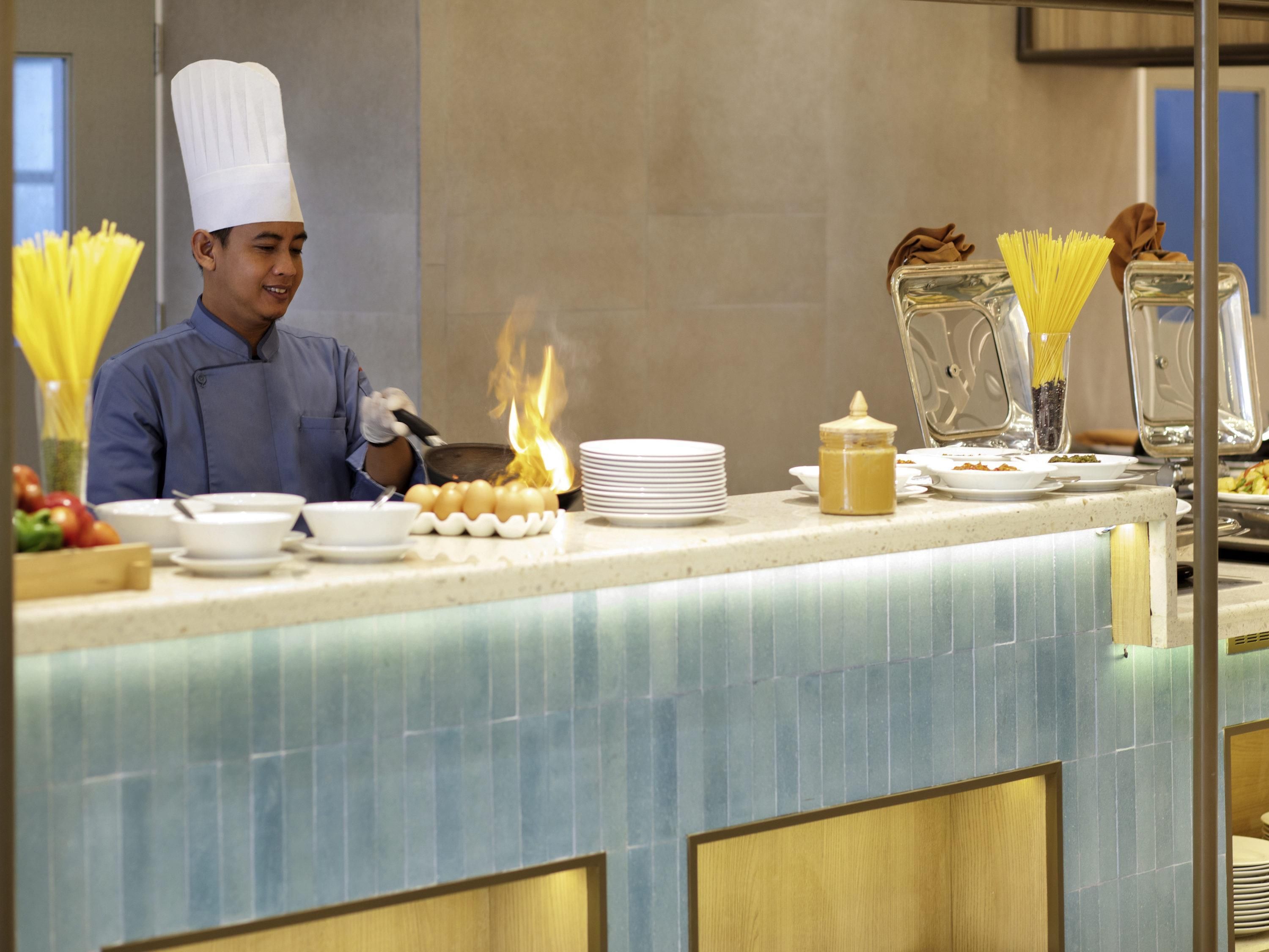 A chef in uniform cooking with visible flames, a buffet counter with plates and bowls, and a tiled backsplash.