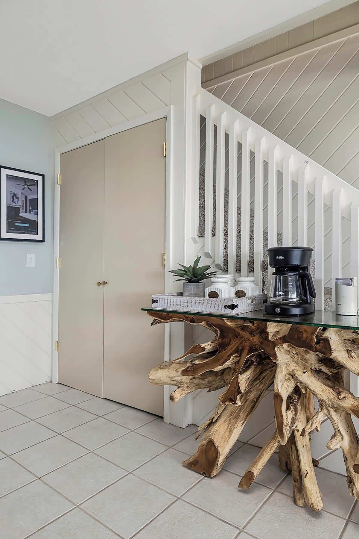 A dining area with a glass-top table featuring a natural wood base, a coffee maker, and a potted plant.