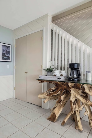 A dining area with a glass-top table featuring a natural wood base, a coffee maker, and a potted plant. - Oceanfront, Pool, GolfCart, & Free EV Charger! (Ocean Isle Beach)