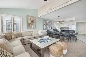 A modern living room with a sectional sofa, a coffee table, and a dining area with chairs. - Oceanfront, Pool, GolfCart, & Free EV Charger! (Ocean Isle Beach)