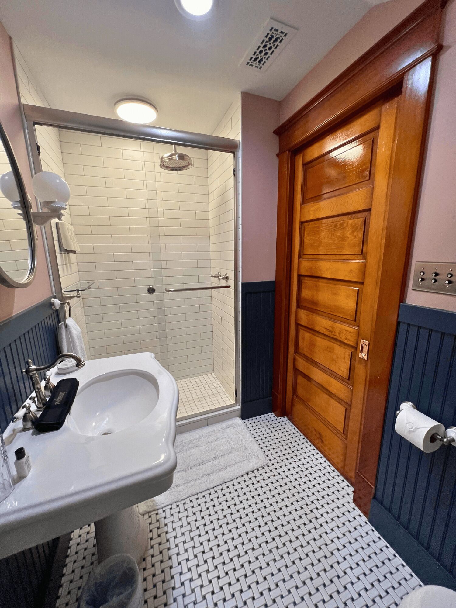 Un baño con un lavamanos blanco, una cabina de ducha de vidrio, una puerta de madera y un suelo con diseño.