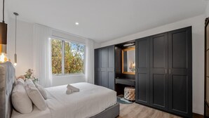 A modern bedroom with a large bed, built-in wardrobe, and a window with a view of trees. - StayCentral - Prahran Penthouse - Mount St, Prahran - 2 Bedrooms, 2 Beds, 2 Bathrooms, 1 secure parking spot and a permit for street parking. (Prahran)