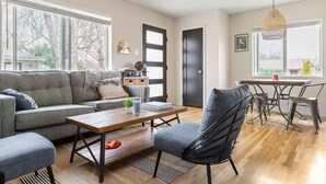 Ein modernes Wohnzimmer mit einer Couch, einem Couchtisch und Sesseln. Es gibt einen Esstisch mit Stühlen, eine Pendelleuchte und eine Tür mit Fenster.