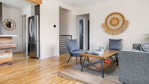 Ein modernes Wohnzimmer mit einem Holzesstisch, zwei grauen Stühlen, einer Küche mit einem Edelstahlkühlschrank und einem Wandbehang.