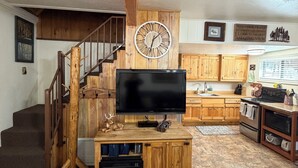 A rustic interior with a wooden staircase, a mounted clock, a flat-screen TV, wooden cabinets, and a kitchen area. - Bell's Greer Hideaway (Greer)