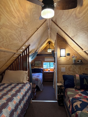 A cozy attic bedroom with a sloped ceiling, a ceiling fan, a window with a view, and a small desk area.