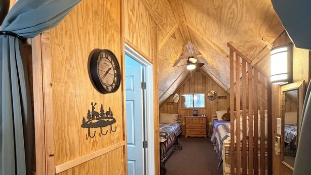 A wooden interior with a clock, a bed, a dresser, and a staircase leading to an upper level. - Bell's Greer Hideaway (Greer)