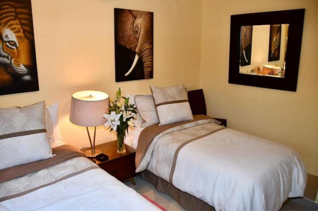Quarto de hotel com duas camas, mesa de cabeceira com abajur e obras de arte nas paredes.