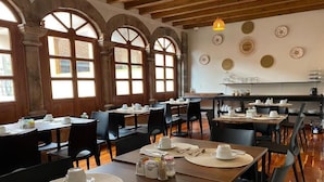 Un espace repas avec plusieurs tables préparées pour le petit-déjeuner, comprenant des tables en bois, des chaises noires et un intérieur rustique avec des poutres apparentes et de grandes fenêtres.
