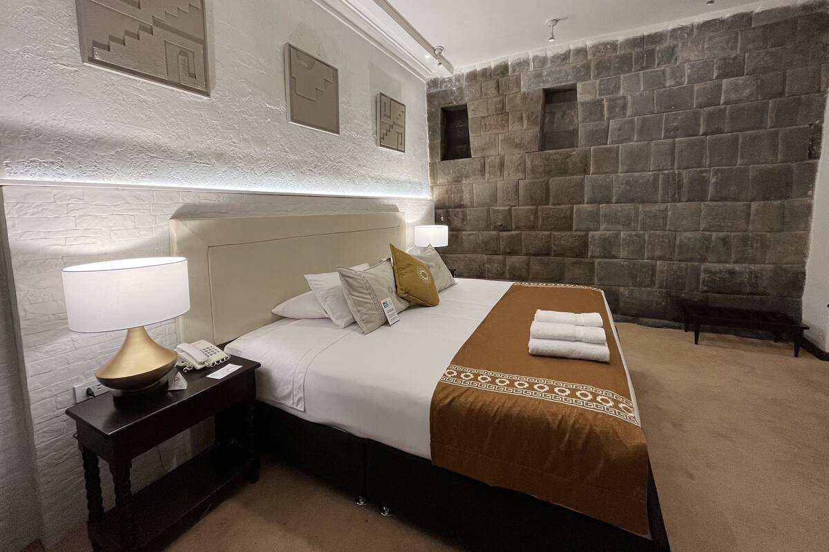A hotel room with a large bed, a nightstand with a lamp, a stone wall, and a small window.
