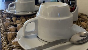Une table avec deux tasses blanches posées sur des soucoupes, une cuillère, une dosette de café Nescafé Dolce Gusto, une bouilloire électrique noire et un verre d’eau.