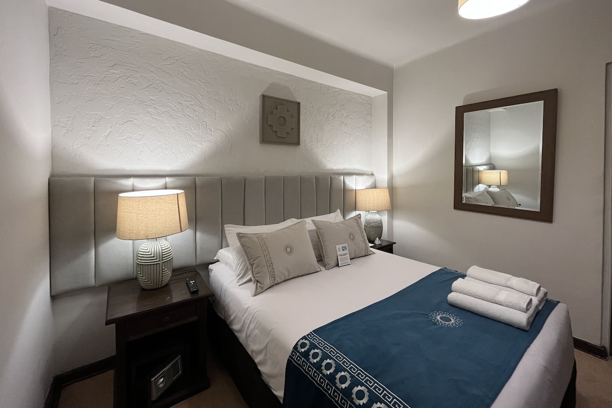 A hotel room with a bed, two bedside tables, a lamp, a mirror, and a wall-mounted picture.