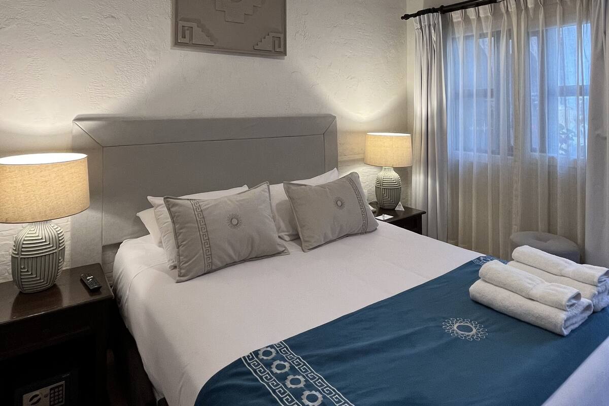 A hotel room with a neatly made bed, a bedside table with a lamp, a wall-mounted artwork, and a window with curtains.