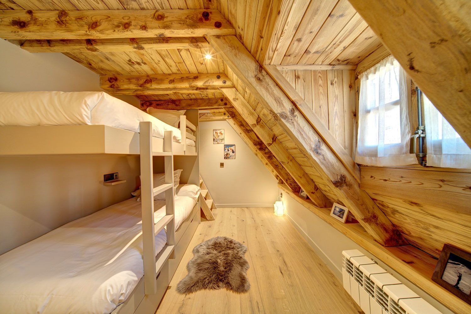 A cozy attic bedroom with wooden beams, bunk beds, a fur rug, and a window with curtains.