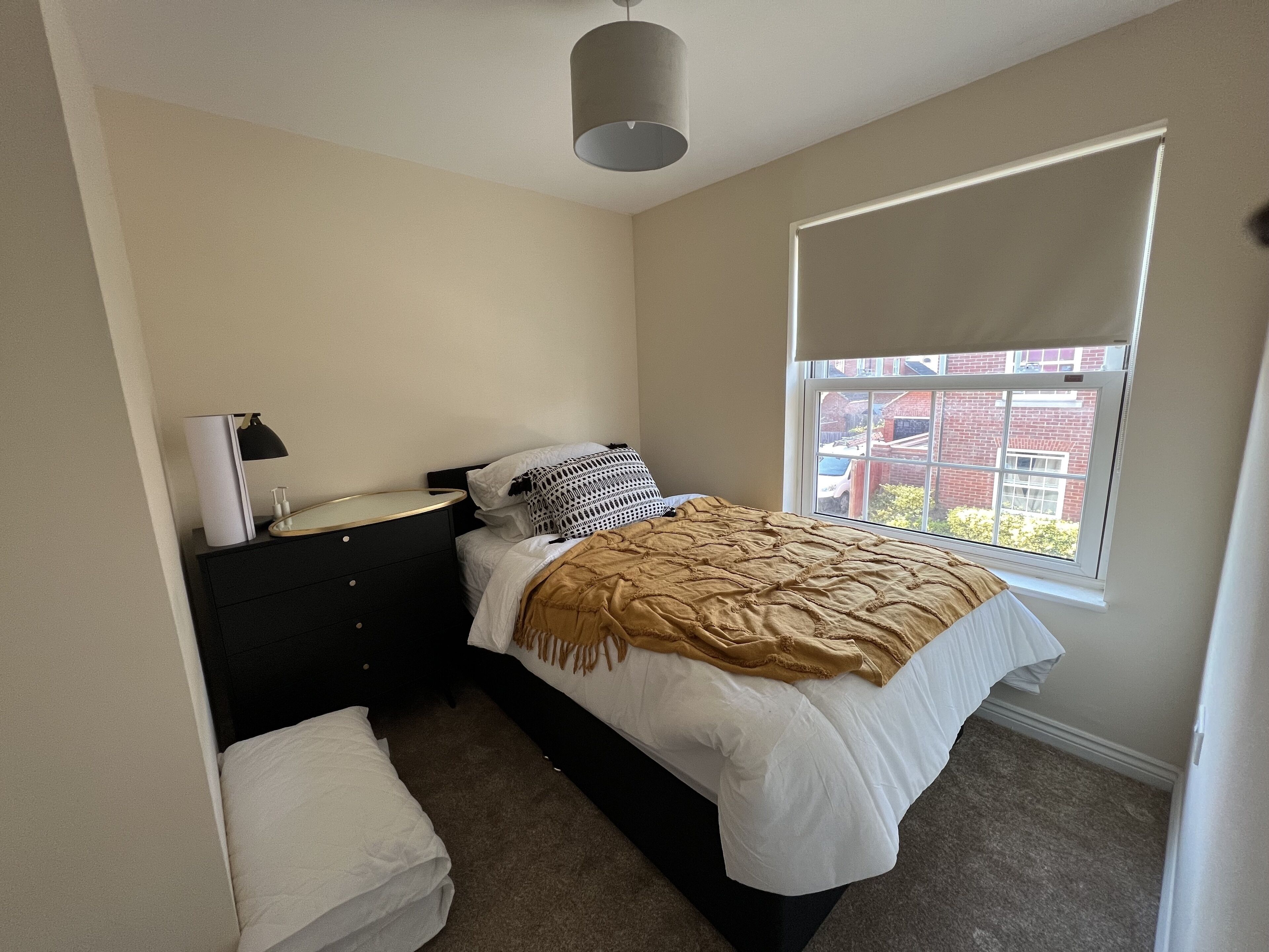 A bedroom with a bed, bedside table, lamp, and a window with a view of buildings.