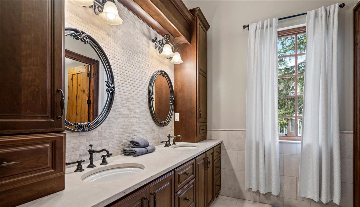 Un baño con dos lavamanos, armarios de madera, un espejo grande y una ventana con cortinas.