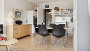 A modern kitchen with white cabinets, a dining area with black chairs, and a small table with a bowl of fruit. - 2 bedroom nice apartment in Fanø (Fanø)