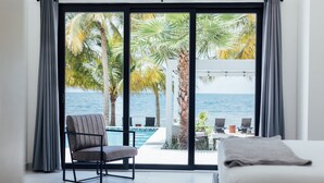 Un dormitorio con un gran ventanal corredera que da a un balcón con piscina y palmeras, con vistas al océano.