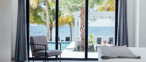 A bedroom with a large sliding glass door leading to a balcony with a pool and palm trees, overlooking the ocean.