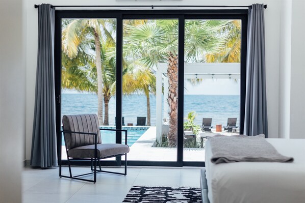Un dormitorio con un gran ventanal corredera que da a un balcón con piscina y palmeras, con vistas al océano.