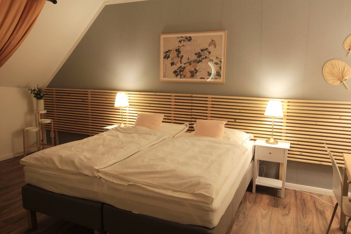 A neatly made bed with a wooden headboard, two bedside tables with lamps, a framed artwork on the wall, and a small plant on the left side.