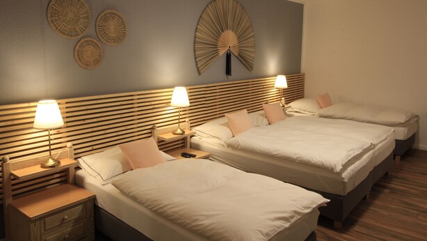 A hotel room with three beds, wooden headboards, bedside tables with lamps, and decorative wall art. - Asia Dreieck Pension Zimmer (Lauterecken)