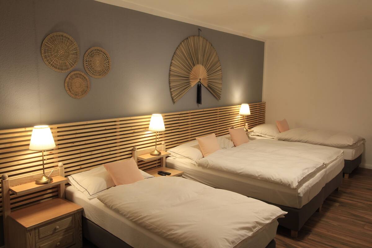 A hotel room with three beds, wooden headboards, bedside tables with lamps, and decorative wall art.