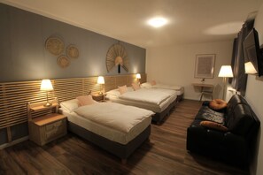 Quarto de hotel com duas camas, um sofá de couro preto, cabeceiras de madeira e luminárias fixadas na parede.