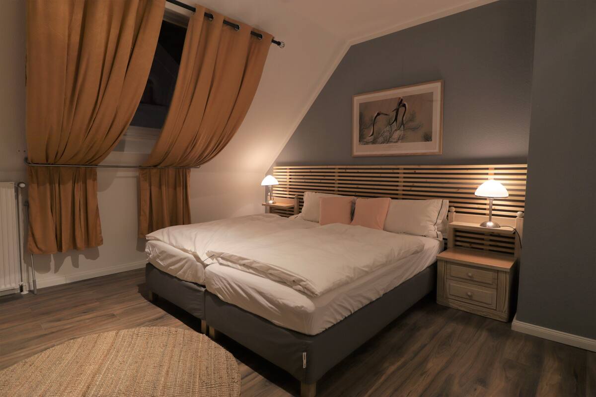 A bedroom with a bed, two bedside tables with lamps, a radiator, and a window with curtains.