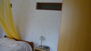 A bedroom with a bed, a nightstand, a lamp, and a window with a curtain. - Maison en rez de Jardin, à 10 Minutes de la mer (Le Morne-Vert)