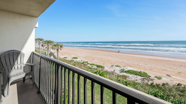 A balcony with a view of a beach, a chair, and a railing. - Oceanfront Charmer (Ormond Beach)
