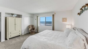 A bedroom with a bed, a chair, a wardrobe, a window with a view, and a lamp. - Oceanfront Charmer (Ormond Beach)